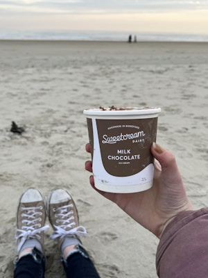 A person holds a cup of Sweetcream Dairy milk chocolate ice cream with a sandy beach in the background. The person is wearing casual shoes and sitting on the sand. The ocean is visible in the distance under a cloudy sky, and two figures are walking along the shore.