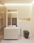 A modern bathroom features a minimalist white vanity with a built-in sink, accompanied by various toiletries arranged neatly. A sleek black toilet contrasts with the neutral tones. A decorative pattern covers the floor tiles, while a black heated towel rail is mounted on the wall. A recessed shelf above the toilet contains an assortment of bottles. Warm ambient lighting enhances the clean and contemporary aesthetic.