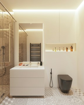 A modern bathroom features a minimalist white vanity with a built-in sink, accompanied by various toiletries arranged neatly. A sleek black toilet contrasts with the neutral tones. A decorative pattern covers the floor tiles, while a black heated towel rail is mounted on the wall. A recessed shelf above the toilet contains an assortment of bottles. Warm ambient lighting enhances the clean and contemporary aesthetic.
