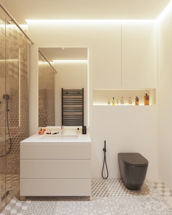 A modern bathroom features a minimalist white vanity with a built-in sink, accompanied by various toiletries arranged neatly. A sleek black toilet contrasts with the neutral tones. A decorative pattern covers the floor tiles, while a black heated towel rail is mounted on the wall. A recessed shelf above the toilet contains an assortment of bottles. Warm ambient lighting enhances the clean and contemporary aesthetic.