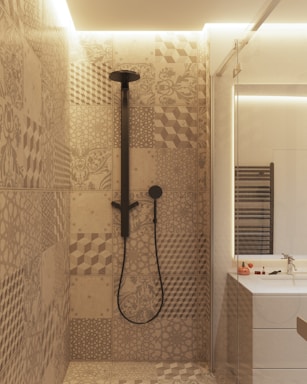 A modern bathroom interior featuring a shower area with intricately patterned tiles in earthy tones. The showerhead and fittings are matte black, adding a contemporary flair. Adjacent to the shower is a vanity with a mirrored section, a sink, and small toiletries placed on the counter. Soft ambient lighting adds a cozy atmosphere to the space.