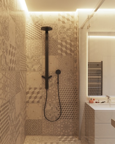 A modern bathroom interior featuring a shower area with intricately patterned tiles in earthy tones. The showerhead and fittings are matte black, adding a contemporary flair. Adjacent to the shower is a vanity with a mirrored section, a sink, and small toiletries placed on the counter. Soft ambient lighting adds a cozy atmosphere to the space.