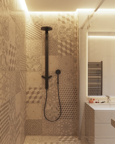 A modern bathroom interior featuring a shower area with intricately patterned tiles in earthy tones. The showerhead and fittings are matte black, adding a contemporary flair. Adjacent to the shower is a vanity with a mirrored section, a sink, and small toiletries placed on the counter. Soft ambient lighting adds a cozy atmosphere to the space.