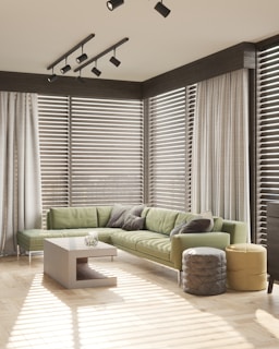 A modern living room with a green L-shaped sofa adorned with gray and white pillows. A contemporary coffee table stands in the center, surrounded by stylish poufs in gray and yellow. Large windows with horizontal blinds allow natural light to fill the room, casting striped shadows on the wooden floor. Ceiling track lights provide additional illumination, complementing the room's minimalist, airy design.