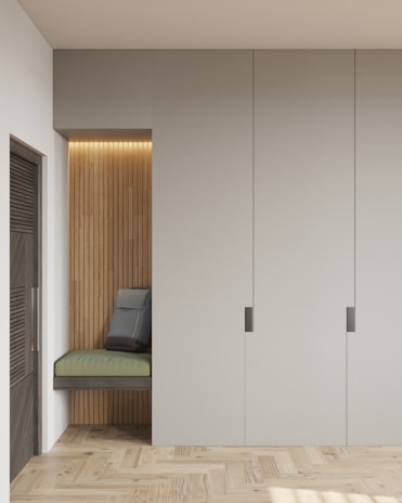 A minimalist interior featuring a built-in seating nook with vertical wooden panels and a soft green cushion. The nook also includes a gray cushion positioned against the wall. Adjacent to the seating area is a modern, smooth-surfaced wardrobe with three large panels and sleek handles. The floor is made of light-colored, textured wood, and soft lighting illuminates the nook.