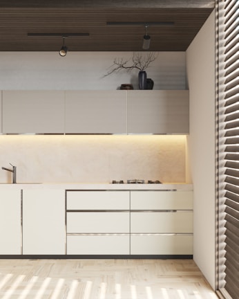 A modern kitchen with light-colored cabinets and sleek design. There are overhead cabinets, a built-in stove, and a minimalist sink. A decorative vase with branches and some pottery are placed on top of the cabinets. The ceiling features two spotlights, and there is ambient lighting under the cabinets. The floor is wooden, and the room is partially lit by sunlight coming through blinds.