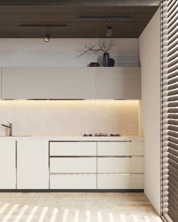 A modern kitchen with light-colored cabinets and sleek design. There are overhead cabinets, a built-in stove, and a minimalist sink. A decorative vase with branches and some pottery are placed on top of the cabinets. The ceiling features two spotlights, and there is ambient lighting under the cabinets. The floor is wooden, and the room is partially lit by sunlight coming through blinds.