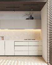 A modern kitchen with light-colored cabinets and sleek design. There are overhead cabinets, a built-in stove, and a minimalist sink. A decorative vase with branches and some pottery are placed on top of the cabinets. The ceiling features two spotlights, and there is ambient lighting under the cabinets. The floor is wooden, and the room is partially lit by sunlight coming through blinds.
