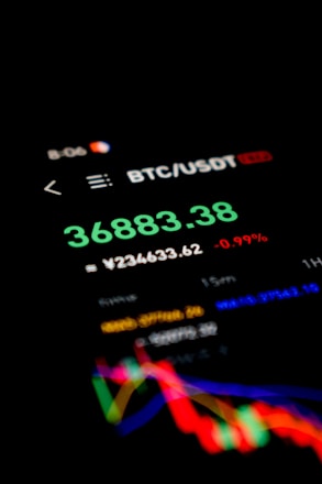 A screen displays cryptocurrency trading information with a focus on the BTC/USDT trading pair. The bold green numbers represent a price of 36883.38, accompanied by a percentage change of -0.99% in red. Below, there is another currency conversion in smaller text and colorful line graphs indicating market trends over time.