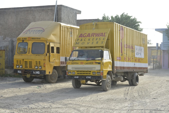 Two yellow moving trucks are parked on a dirt road next to a building. The trucks are labeled with 'Agarwal Packers & Movers,' indicating they are used for transporting goods. The background includes a simple building with minimal decoration and some greenery.