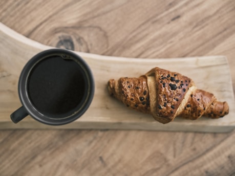 A freshly baked croissant with a flaky, golden-brown crust topped with chocolate chips lies on a wooden serving board. Next to it, a black ceramic cup filled with dark coffee sits, creating an inviting and cozy breakfast scene.