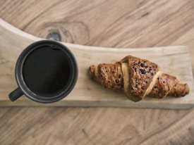 A freshly baked croissant with a flaky, golden-brown crust topped with chocolate chips lies on a wooden serving board. Next to it, a black ceramic cup filled with dark coffee sits, creating an inviting and cozy breakfast scene.