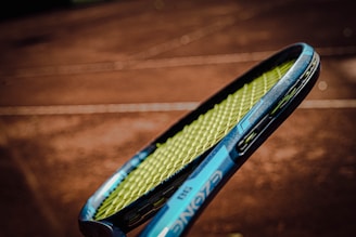 A close-up of a tennis racket with a blue frame and yellow strings, held against the blurred background of a clay tennis court.