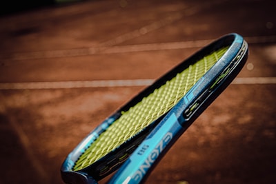 A close-up of a tennis racket with a blue frame and yellow strings, held against the blurred background of a clay tennis court.