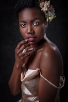 A woman with short curly hair adorned with a floral arrangement, wearing a satin beige dress. She is posed with her hand gently resting on her chin, exhibiting a confident expression. The lighting highlights her smooth skin and elegant posture against a dark backdrop.