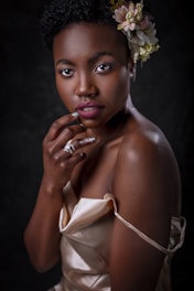 A woman with short curly hair adorned with a floral arrangement, wearing a satin beige dress. She is posed with her hand gently resting on her chin, exhibiting a confident expression. The lighting highlights her smooth skin and elegant posture against a dark backdrop.