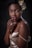 A woman with short curly hair adorned with a floral arrangement, wearing a satin beige dress. She is posed with her hand gently resting on her chin, exhibiting a confident expression. The lighting highlights her smooth skin and elegant posture against a dark backdrop.