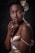 A woman with short curly hair adorned with a floral arrangement, wearing a satin beige dress. She is posed with her hand gently resting on her chin, exhibiting a confident expression. The lighting highlights her smooth skin and elegant posture against a dark backdrop.