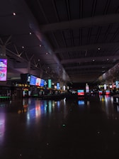 An expansive indoor space with high ceilings and numerous illuminated digital screens displaying various advertisements and information. The polished floor reflects the colorful lights, and the area appears spacious and quiet.