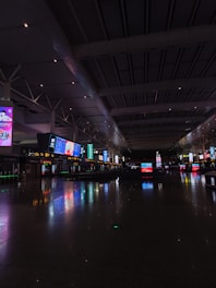 An expansive indoor space with high ceilings and numerous illuminated digital screens displaying various advertisements and information. The polished floor reflects the colorful lights, and the area appears spacious and quiet.