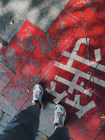 A pair of feet in white sneakers standing on a red and gray textured pavement with faded white text. The ground appears worn and weathered, with a dry leaf on one side, suggesting an outdoor urban setting.