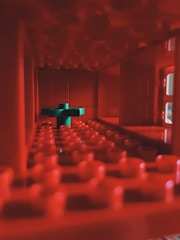 A close-up view inside a structure made of red interlocking plastic bricks, possibly LEGO. In the center, a contrasting green brick is placed, creating a focal point amidst the monochromatic red tones. The lighting accentuates the texture and shadows within the cavity.