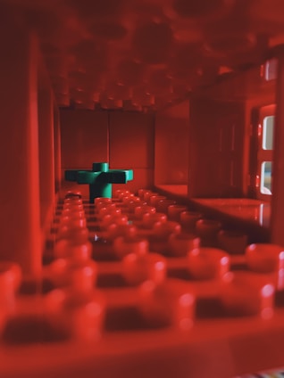 A close-up view inside a structure made of red interlocking plastic bricks, possibly LEGO. In the center, a contrasting green brick is placed, creating a focal point amidst the monochromatic red tones. The lighting accentuates the texture and shadows within the cavity.