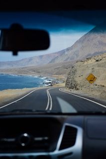 The image captures a scenic coastal road as viewed from the inside of a vehicle, looking through the windshield. The road winds along the coastline with the ocean on one side and rugged hills on the other. A yellow traffic sign is visible on the right, and the sky is clear with a few clouds.