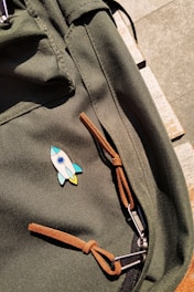 A close-up of a green fabric backpack featuring a colorful rocket-shaped pin. The bag has brown leather straps and silver zippers set against a light wooden surface.