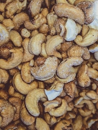 A close-up view of numerous roasted cashew nuts with visible salt crystals on their surfaces. The nuts are irregularly shaped, with a warm, golden-brown hue, and some pieces show a glossy roasted texture.