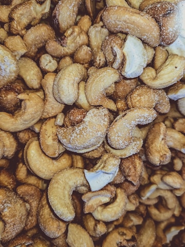 A close-up view of numerous roasted cashew nuts with visible salt crystals on their surfaces. The nuts are irregularly shaped, with a warm, golden-brown hue, and some pieces show a glossy roasted texture.