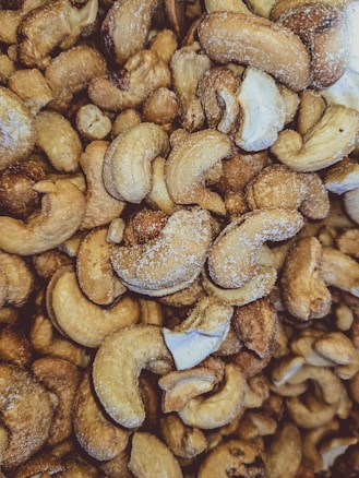 A close-up view of numerous roasted cashew nuts with visible salt crystals on their surfaces. The nuts are irregularly shaped, with a warm, golden-brown hue, and some pieces show a glossy roasted texture.