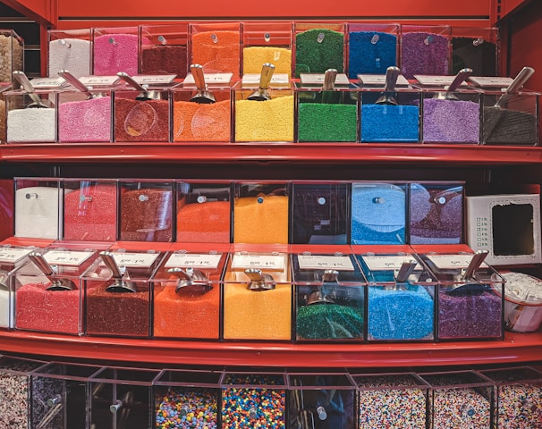 An assortment of colorful sprinkles and toppings displayed in clear containers on red shelves. Each container is filled with a different color, including pink, orange, yellow, green, blue, purple, and black. Small scoops are placed in each container for customers to use.
