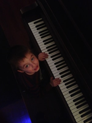 A young child is looking up while standing in front of a piano keyboard. The piano is positioned diagonally, and the lighting is dim with a bluish hue on the floor. The child has brown hair and is wearing a dark striped shirt.