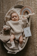 A baby wearing a beige knitted sweater and white socks is lying in a woven basket on a textured carpet. Next to the baby is a colorful, arched toy rainbow made of fabric and a card that says '3 Months Old'. The baby is looking at the camera with a calm expression.