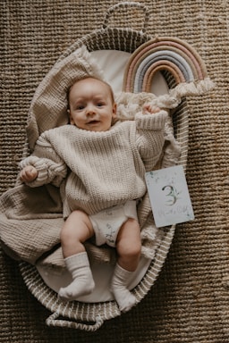 A baby wearing a beige knitted sweater and white socks is lying in a woven basket on a textured carpet. Next to the baby is a colorful, arched toy rainbow made of fabric and a card that says '3 Months Old'. The baby is looking at the camera with a calm expression.