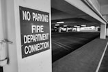 A black and white image of an empty parking garage features a prominently displayed sign on a pillar that reads 'No Parking Fire Department Connection.' The garage has several visible parking lines, ceiling lights, and is devoid of vehicles or people, giving it a deserted appearance.