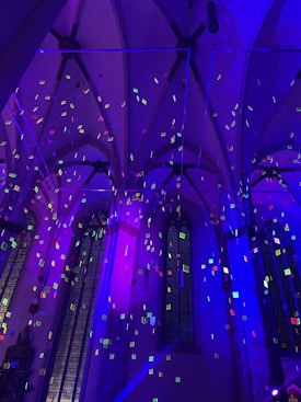 Gothic-style architecture with high arches and stained glass windows illuminated by vivid purple and blue lighting. Numerous small, colorful squares hover in the air, contributing to a surreal and dynamic atmosphere.