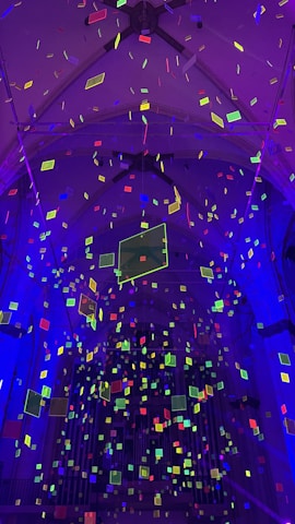 An indoor scene with architectural elements illuminated by a vibrant display of colorful rectangular shapes suspended in the air. The shapes are primarily neon green, yellow, pink, and blue, creating a dynamic and futuristic atmosphere against a backdrop of dark and arched ceiling structures.