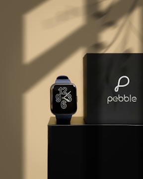 A smartwatch with a modern design sits elegantly on a glossy black surface. The watch face displays large, stylized numbers in white. Adjacent to the watch is a black box with the brand name 'pebble' and its logo printed in white. The background is softly illuminated with shadow patterns that add depth and contrast.