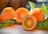 Fresh oranges are displayed on a light wooden surface, with one orange cut in half to reveal its juicy interior. Green leaves are attached to the oranges, adding a natural and vibrant touch.