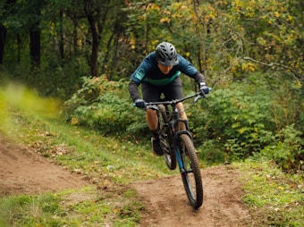 A person is mountain biking on a dirt trail surrounded by lush greenery and trees. The person is wearing a helmet and protective gear, and is focused and leaning forward on the bike, navigating through the path.