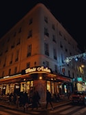 A corner cafe with illuminated signage reading 'Paris Auber' sits on a bustling city street at night. People walk along the sidewalk, and patrons dine at tables outside the brasserie. The building is adorned with festive lights, and a car is parked nearby.