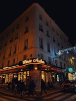A corner cafe with illuminated signage reading 'Paris Auber' sits on a bustling city street at night. People walk along the sidewalk, and patrons dine at tables outside the brasserie. The building is adorned with festive lights, and a car is parked nearby.