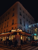 A corner cafe with illuminated signage reading 'Paris Auber' sits on a bustling city street at night. People walk along the sidewalk, and patrons dine at tables outside the brasserie. The building is adorned with festive lights, and a car is parked nearby.
