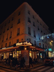 A corner cafe with illuminated signage reading 'Paris Auber' sits on a bustling city street at night. People walk along the sidewalk, and patrons dine at tables outside the brasserie. The building is adorned with festive lights, and a car is parked nearby.