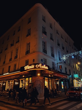 A corner cafe with illuminated signage reading 'Paris Auber' sits on a bustling city street at night. People walk along the sidewalk, and patrons dine at tables outside the brasserie. The building is adorned with festive lights, and a car is parked nearby.