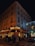 A corner cafe with illuminated signage reading 'Paris Auber' sits on a bustling city street at night. People walk along the sidewalk, and patrons dine at tables outside the brasserie. The building is adorned with festive lights, and a car is parked nearby.