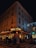 A corner cafe with illuminated signage reading 'Paris Auber' sits on a bustling city street at night. People walk along the sidewalk, and patrons dine at tables outside the brasserie. The building is adorned with festive lights, and a car is parked nearby.
