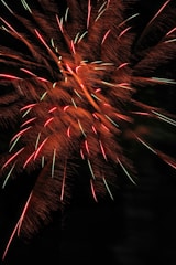 Bright, colorful fireworks burst against a dark night sky with vibrant streaks and patterns of red, orange, and hints of green, creating an explosive display.